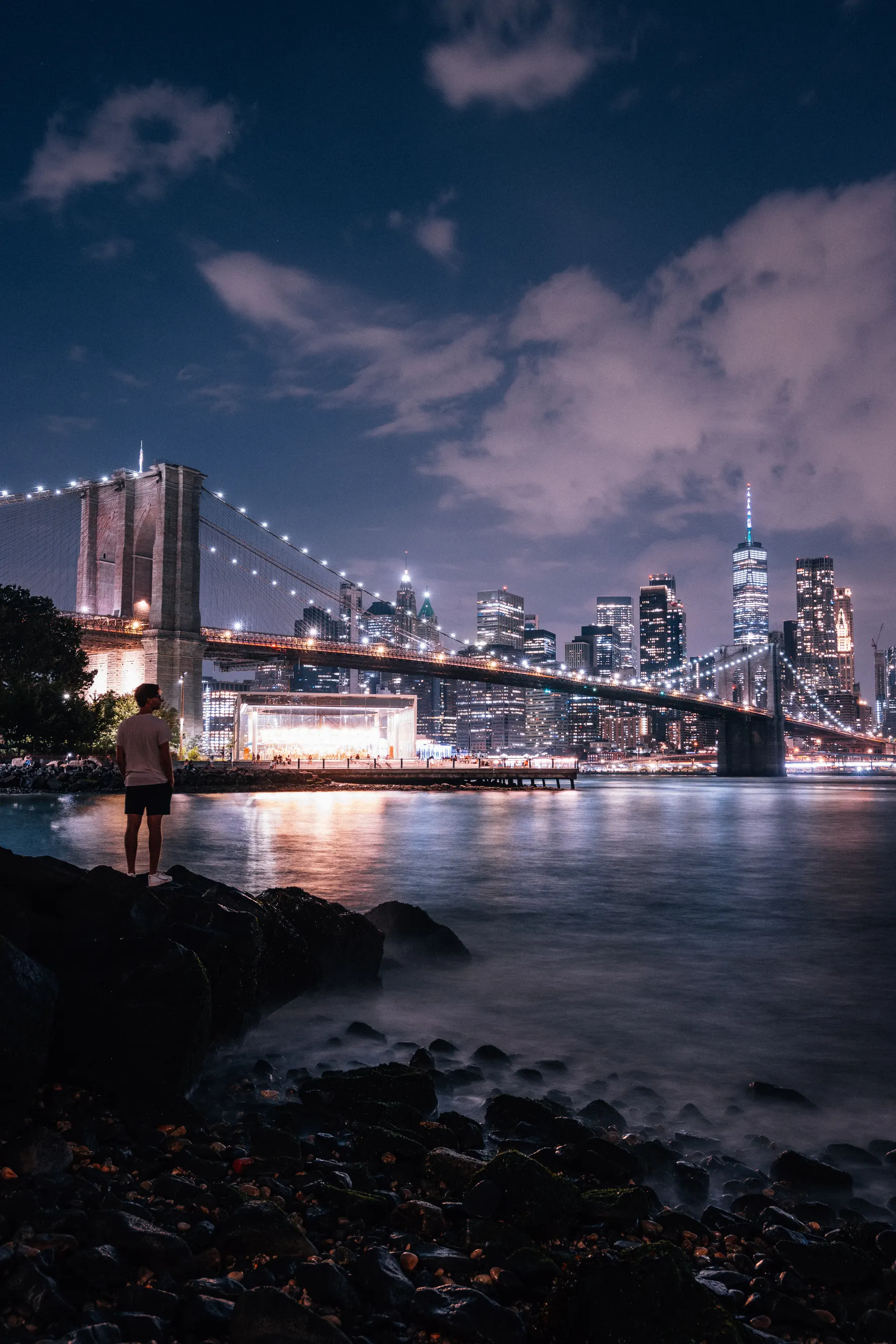 The lit bridge and the Manhattan skyline at night, seen across the water from the rocks