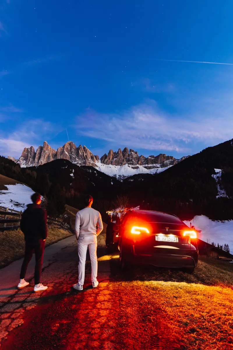 Jagged snow-covered peaks under a deep blue night sky, tail lights on the road below