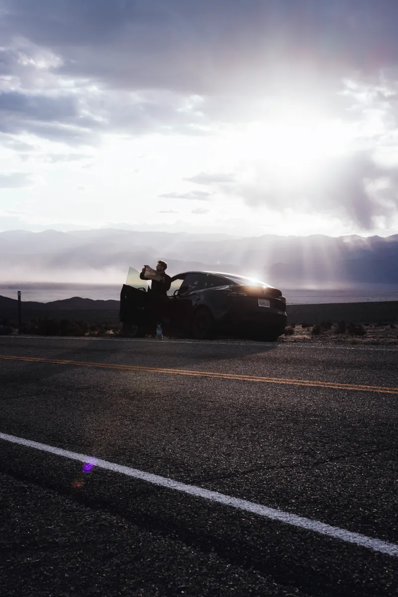 Car stopped on an empty desert road, backlit by low sun over distant mountains